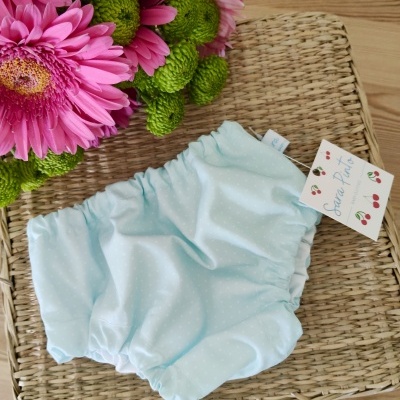 Cuecas para bebé azul claro com bolinhas brancas e etiqueta em superfície de palha com flores.