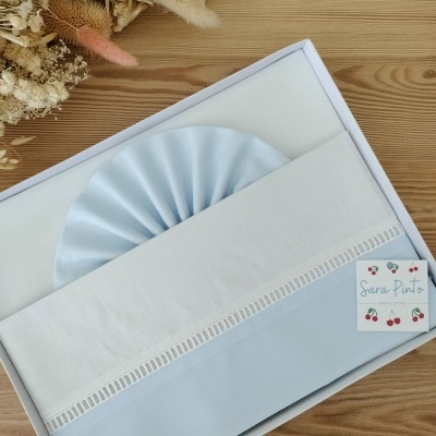 Conjunto de roupa de cama azul e branca em caixa com etiqueta Sara Pinto e flores secas ao lado