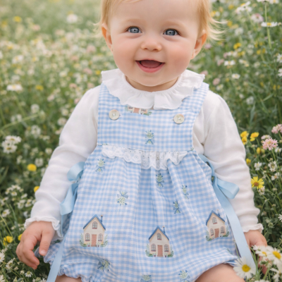 Bebé loiro sentado numa campo florido com roupa azul clara xadrez com casas bordadas.
