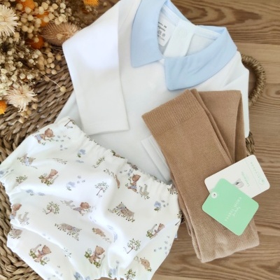 Conjunto de roupa de bebé com body branco com gola azul, calção branco com padrão de desenhos e meias castanhas.