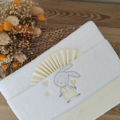 Toalha branca com bordado de coelho e corações em cinza e amarelo sobre mesa de madeira com flores secas.