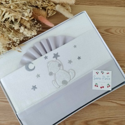 Lençol de berço branco e cinzento com bordado de ursinho e estrelas em caixa branca ao lado de ramo seco