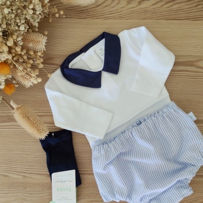 Conjunto de roupa de bebé com camisa branca e gola azul escura, calções às riscas azuis e brancos, meias azuis escuras, sobre madeira clara com flores secas.