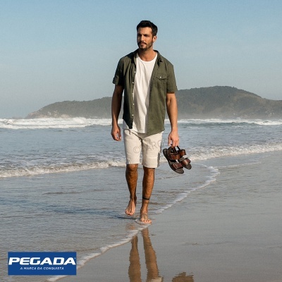 Homem com roupa casual a caminhar na praia, segurando sandálias.