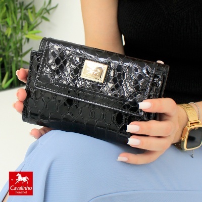 Carteira preta brilhante com padrão crocodilo e fecho dourado sendo segurada por uma pessoa