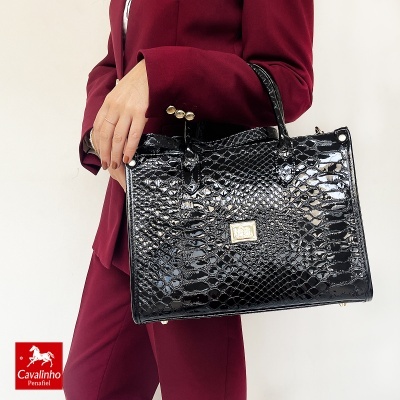 Mala de mão preta texturizada brilhante com alças e placa metálica dourada, segura por pessoa de fato bordeaux