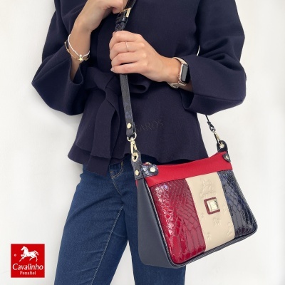 Mala senhora Cavalinho com padrão pele cobra vermelho, creme e azul escuro, alça ajustável, vista num corpo com camisa preta e jeans.