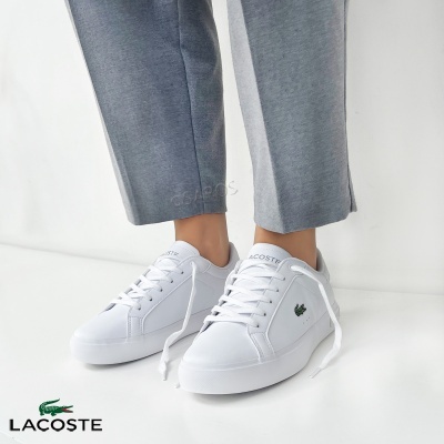 Sapatilhas brancas Lacoste com logótipo crocodilo verde e calças cinzentas em fundo branco