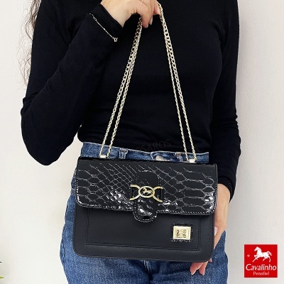 Mala preta com padrão texturizado e fecho dourado, segurada por uma mulher com roupa preta e jeans azuis