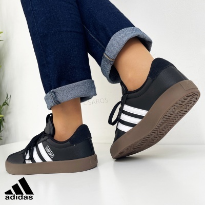 Tênis Adidas pretos com riscas brancas e sola castanha usados com calças de ganga