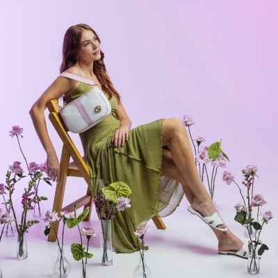 Mulher sentada com vestido verde, bolsa branca e sandálias brancas rodeada de flores em vasos de vidro