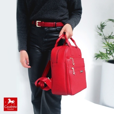 Mochila vermelha de material sintético com fechos dourados e alças, usada por pessoa com cinto vermelho e roupa cinza e preta