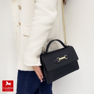 Mala preta de pele com alça dourada e fecho metálico, segurada por pessoa com casaco branco e calças de ganga