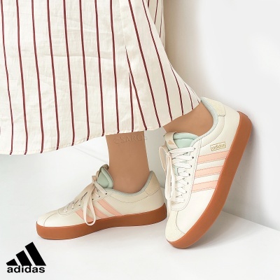 Ténis Adidas brancos com riscas cor de rosa e sola castanha, calçado por pessoa com roupa listrada.