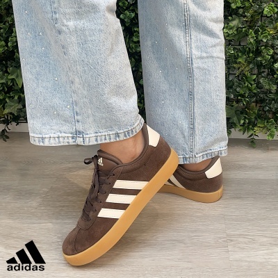 Ténis Adidas castanhos com riscas brancas e sola castanha claramente visíveis com calças de ganga e planta de fundo