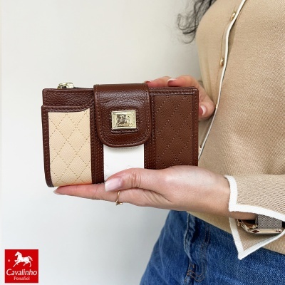 Carteira feminina Cavalinho em couro castanho e tecido bege com logótipo dourado, segurada pela mão de pessoa com camisola bege e jeans