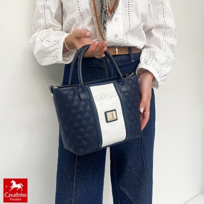 Mala pequena de senhora em pele azul escuro com faixa branca e emblema, segurada por pessoa com camisa branca bordada e calças de ganga.