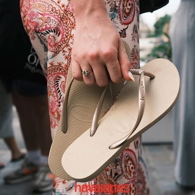 Chinelos Havaianas bege com tiras douradas em mão feminina junto a calças com padrão colorido