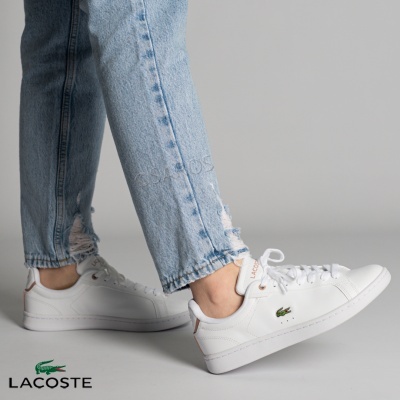 Ténis Lacoste brancos com logo de crocodilo verde e detalhe castanho, usados com calças de ganga claras rasgadas