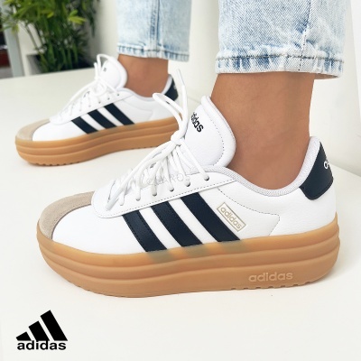 Ténis Adidas brancos com riscas pretas e sola castanha, usados por uma pessoa