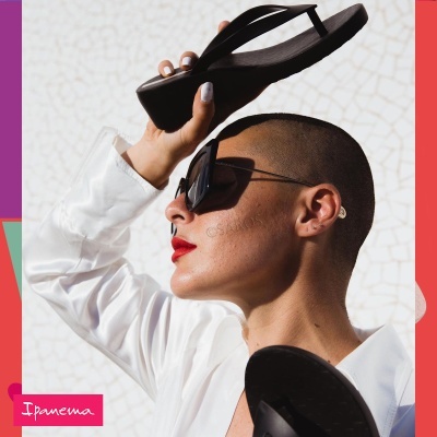 Pessoa com óculos escuros segurando chinelos pretos de plataforma com camisa branca e batom vermelho