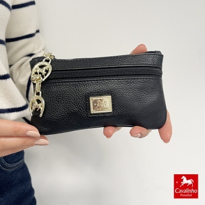 Carteira preta em pele com fechos de correr e pendente dourado, segurada por uma pessoa