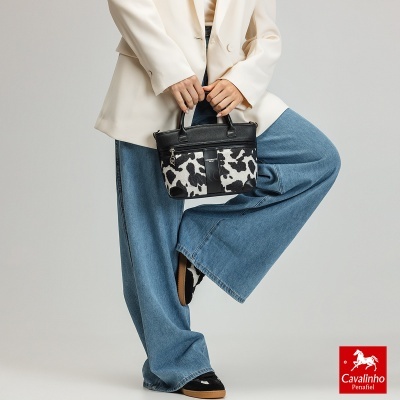 Mala de mão pequena preta com padrão de manchas preto e branco, segurada por pessoa com blazer bege e jeans azuis largos