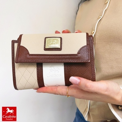 Carteira Cavalinho bege, branco e castanho com logo dourado, sendo segurada por mão feminina