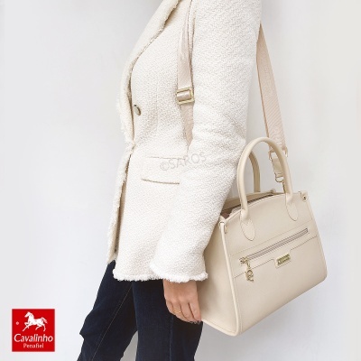 Mala de mão bege com bolso frontal e alça larga ajustável exibida por mulher com casaco branco