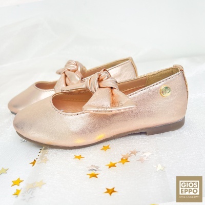 Sapatos infantis dourados com laço e sola castanha sobre fundo branco com estrelas