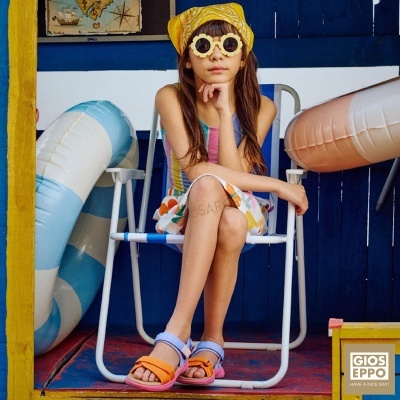 Menina com vestido colorido, chapéu amarelo e óculos de sol sentada numa cadeira branca e azul