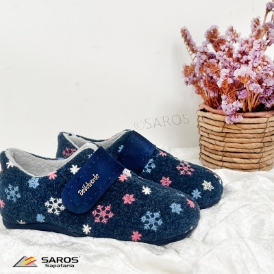 Chinelos azuis com padrão de flocos de neve e fecho de velcro azul, ao lado de um vaso de flores.