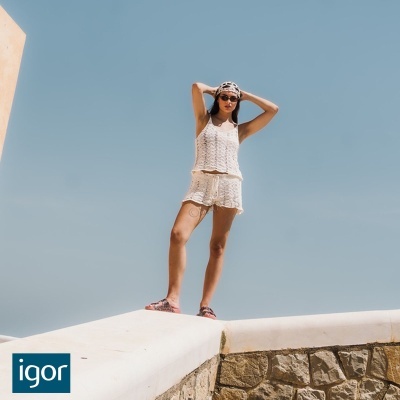 Mulher jovem com conjunto de croché branco, lenço na cabeça, óculos de sol e chinelos, em cenário ao ar livre com céu azul