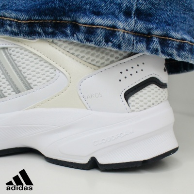 Parte de trás de um tênis branco Adidas com detalhes pretos e calça de ganga azul