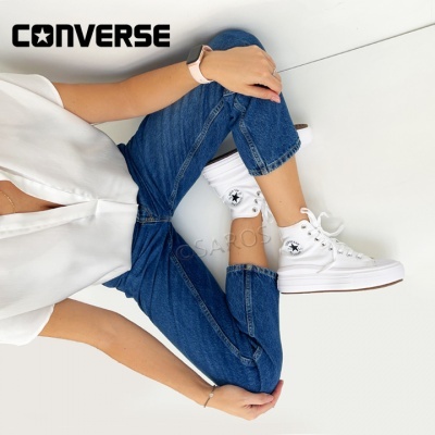 Pessoa a usar ténis Converse brancos sentado no chão com calças de ganga e blusa branca