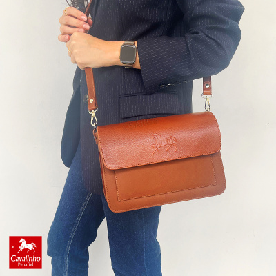 Mala castanho claro em couro com relevo de cavalo na aba, alça ajustável castanha, vestida com blazer azul e jeans azuis