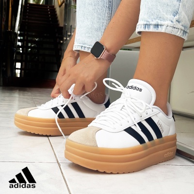 Ténis Adidas brancos com riscas pretas e sola grossa cor de caramelo em piso cerâmico branco