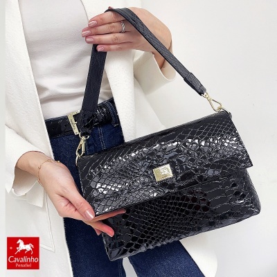 Mala preta de padrão crocodilo com fecho metálico prateado, alça de mão, em fundo claro, segura por pessoa vestindo casaco branco e jeans azul.