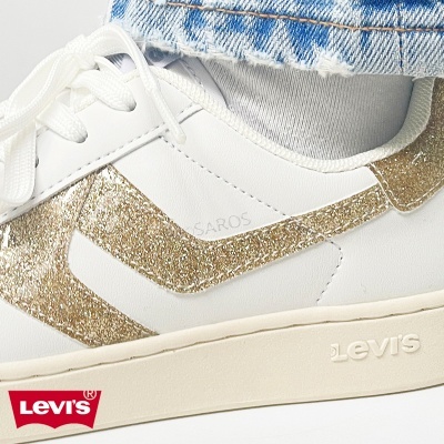 Ténis brancos Levis com detalhes dourados e sola bege, usados com meias brancas e calças de ganga rasgadas.