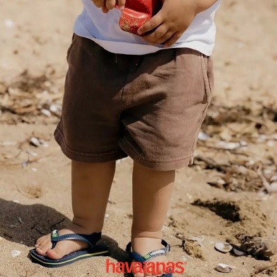 Criança com calções castanhos, t-shirt branca e sandálias Havaianas azul e verde na praia