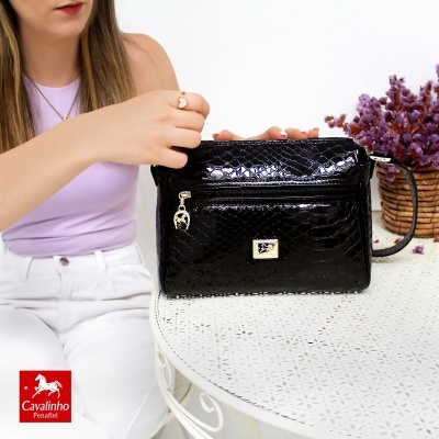 Mala preta com padrão croco numa mesa branca ao lado de vaso com flores lilases