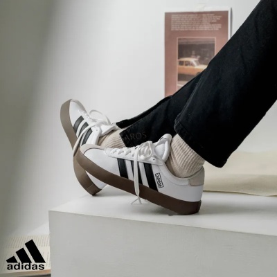 Ténis Adidas brancos com riscas pretas e sola castanha num ambiente interior minimalista.