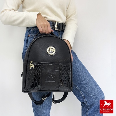 Mochila preta Cavalinho com textura croco e detalhes dourados, segurada por pessoa com camisola creme e jeans azuis