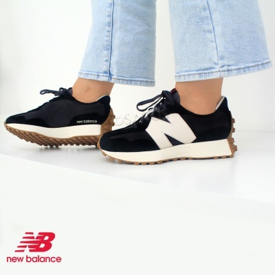 Ténis New Balance pretos e brancos com sola castanha, calçado em ambiente minimalista, com logo New Balance vermelho no canto inferior esquerdo.
