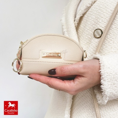 Carteira pequena bege com placa dourada Cavalinho, segurada por mão e casaco bege