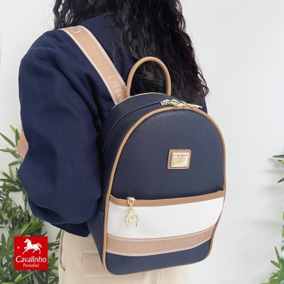 Mochila pequena preta, branca e bege com detalhes dourados e plaqueta frontal, usada por pessoa com casaco azul escuro.