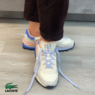 Ténis Lacoste branco, azul, rosa e cinzento com atacadores lilás no chão de madeira