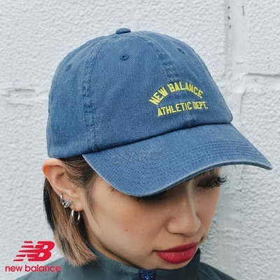 Boné azul com bordado amarelo 'NEW BALANCE ATHLETIC DEPT.' usado por pessoa junto a parede branca