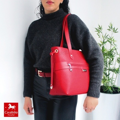 Mala vermelha de pele com fecho zip e alças duplas, sendo segurada por pessoa com camisola cinza escuro e calças pretas