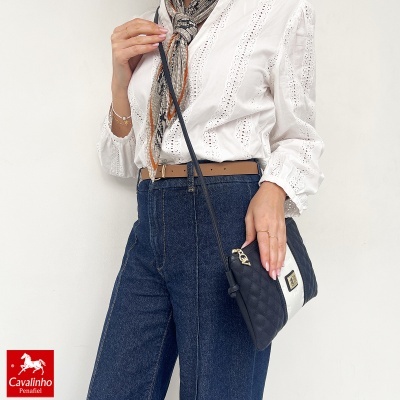 Mala bicolor preta e branca com padrão matelassê e fecho dourado, usada por uma pessoa com camisa branca bordada, jeans azuis e lenço às riscas.